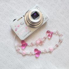 a camera and some pink beads on a white table cloth next to a cell phone