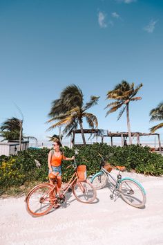 a woman standing next to two bikes on the beach with palm trees in the background