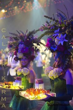 two women dressed in green dresses holding trays with flowers on them and lights behind them