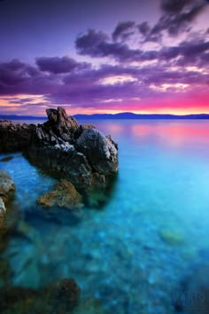 the water is crystal blue and clear with rocks in front of it, as the sun sets