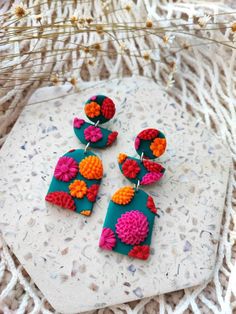 three pairs of colorful earrings sitting on top of a white surface next to dried flowers