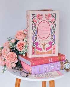 a stack of books sitting on top of a table next to pink flowers and an alarm clock