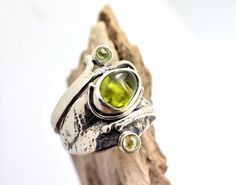 Lovely raw tumbled peridot gemstone and two small round peridots on a freeform leaf band handcrafted from sterling silver The band of the ring it is formed as a leaf and is wrapping the finger ending to small helixes. It is one of a kind ring. The shank of the ring is unusual, textured and have a light oxidation. It is designed on Lesbos island by us Nikiforos and Nelly inspired from walks into the greek nature. The ring is one of a kind and is made in US size 6 ,European 52 and UK M round perid Fine Jewelry With Peridot Gemstone, Nature-inspired Jewelry With Natural Stones In Freeform, Bohemian Sterling Silver May Birthstone Jewelry, Unique Natural Stone May Birthstone Jewelry, Bohemian Sterling Silver Jewelry For May Birthstone, Unique Jewelry With Natural May Birthstones, Unique Natural Stones Jewelry For May Birthstone, Unique Gemstone Jewelry For May Birthstone, Unique May Birthstone Gemstone Jewelry