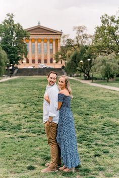 a man and woman standing in front of a building on the grass with trees behind them