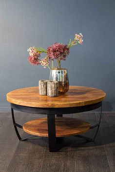 a round wooden table with metal legs and flowers in a vase on the top, against a gray wall