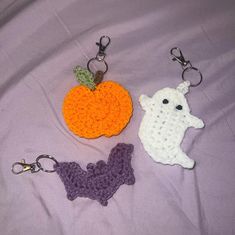 three crocheted keychains are laying on a purple sheet and one has an orange pumpkin, the other is a ghost