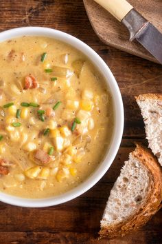 a bowl of corn chowee next to a piece of bread on a wooden table