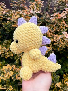 a hand holding a small yellow and purple crocheted stuffed animal in front of some bushes