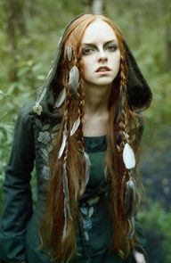 a woman with long red hair and feathers on her head is standing in the woods