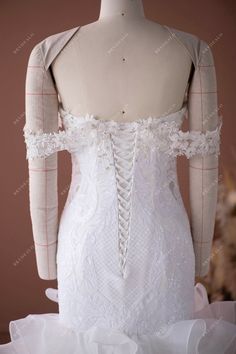 the back of a white wedding dress with lace and ruffles on it, in front of a mannequin