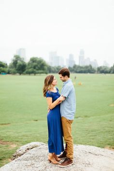 a pregnant couple standing on top of a rock in a field with the city skyline behind them