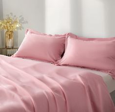 a bed with pink sheets and pillows in a white room next to a flower vase