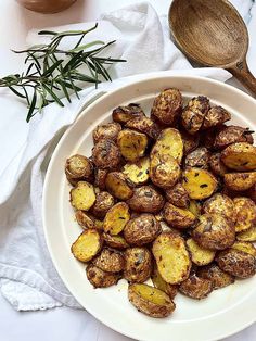 a white plate topped with cooked potatoes next to a wooden spoon and sprig of rosemary