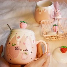 a person holding a pink mug with strawberries on it next to two other cups