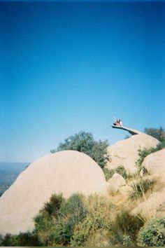 two people are sitting on the top of a large rock with trees in the background