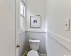 a white toilet sitting next to a window in a bathroom under a framed art piece