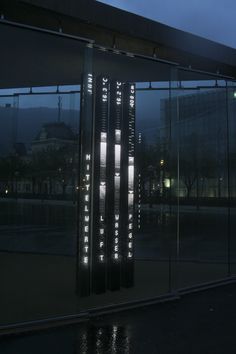 an illuminated clock on the side of a glass building at night with buildings in the background