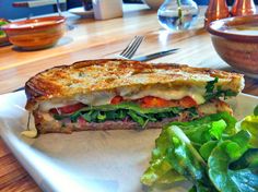 a sandwich with meat and vegetables on a white plate next to a bowl of salad