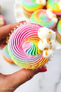 a hand holding a rainbow cupcake with white frosting and sprinkles