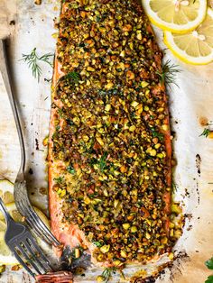 a piece of salmon covered in nuts and herbs next to a fork, lemon wedges and knife