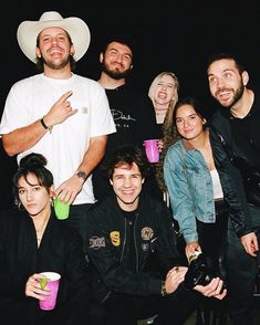 a group of people standing next to each other in front of a black background with one man wearing a cowboy hat