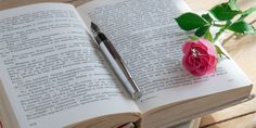 an open book with a pen on top of it next to a pink rose and green leaves