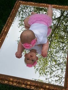 a baby in pink and white outfit standing on grass looking at herself in the mirror