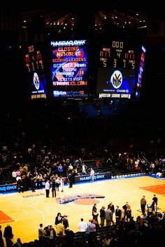 an indoor basketball court with people standing on the sidelines and score boards in the background