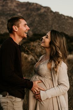 a man and woman standing next to each other in front of a mountain with their arms around each other