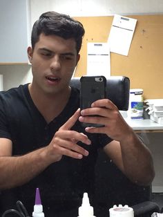 a man taking a selfie with his cell phone in front of him and bottles of nail polish on the table behind him