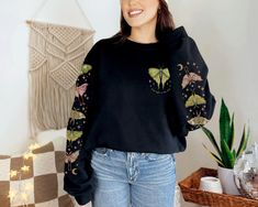 This beautiful Luna Moth sweatshirt with the phrase "find light in the dark" has a gorgeous sleeve print on both sleeves, with dark celestial nature vibes. Our crewneck sweatshirts have a loose fit and are made with a medium-heavy cotton/poly blend making them warm, comfortable, and cozy. There are no itchy side seams and the collars are ribbed knit, so they retain their shape even after washing. SPECIFICATIONS ❤️ Unisex sizing ❤️ Medium-heavy cozy fabric ❤️ Sewn-in label ❤️ Loose fit, size up for an oversized look ❤️ 50% cotton, 50% polyester ❤️ Colors may slightly vary due to monitor differences PRINTING PROCESS We use direct-to-garment printing, a much more sustainable method than older printing methods, like screen printing. We apply pigmented water-based inks that are absorbed by the Aesthetic Long Sleeve Sweatshirt For Fall, Goblincore Sweater, Dark Celestial, Granola Girl Aesthetic, Dark Cottagecore, Aesthetic Nature, Luna Moth, Granola Girl, Sew-in Labels