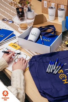 Photo of a person's hands writing gift tags on a desk that is covered in branded items like insulated mugs, notebooks, pens, and tote bags Company Merch, Products Ads, Table Swag, Candy Gift Box, Kit Ideas, Custom Ink, Care Packages