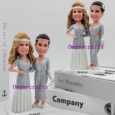 three bobble heads are shown on top of the company's packaging boxes, and one is wearing a silver dress