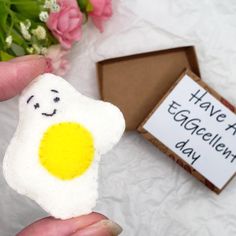 someone is holding up a piece of felt with a smiley face on it next to some flowers