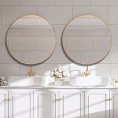 two round mirrors on the wall above sinks in a bathroom with white cabinets and gold trim