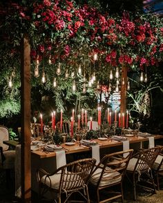 an outdoor dining table with candles and flowers hanging from it's ceiling, surrounded by greenery