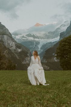 a woman in a white dress is walking through the grass with mountains in the background