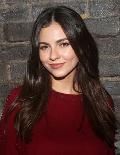 a woman with long brown hair wearing a red sweater and smiling at the camera while standing in front of a brick wall