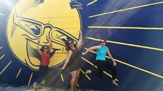 three women are posing in front of a mural on the side of a building that is painted yellow and blue