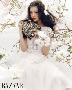 a woman in a white dress holding a purse with flowers around her neck and sitting on the ground