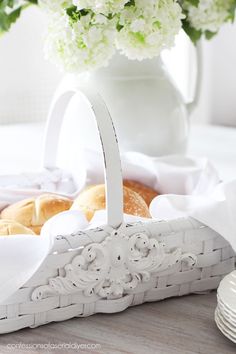 a white basket filled with bread next to a vase full of flowers and plates on a table