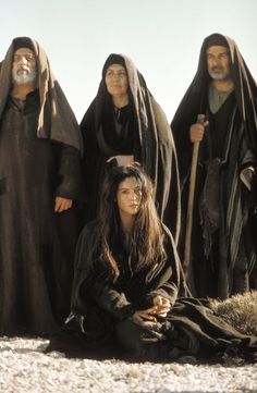 three people dressed in black sitting on the ground with one person holding a stick and two others standing behind them