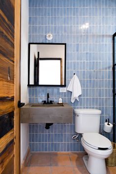 a blue tiled bathroom with a toilet and sink under a mirror in the corner next to a wooden door