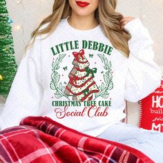 a woman wearing a white shirt with a christmas tree on it and the words, little debie christmas tree cake social club