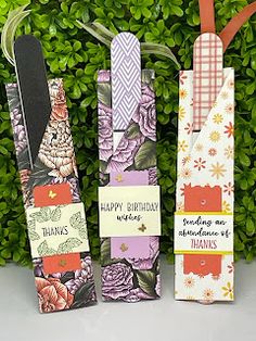 three bookmarks with flowers on them are sitting in front of some green plants and bushes