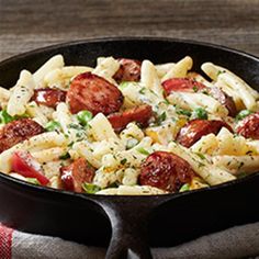 a skillet filled with pasta and sausage on top of a wooden table next to a towel