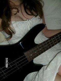 a woman laying in bed with a black electric bass guitar on her lap and looking at the camera