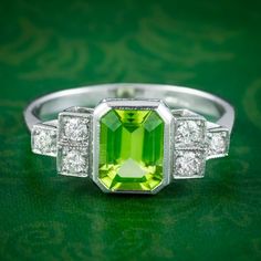 "A striking Art Deco inspired ring fashioned in platinum and bezel set with an emerald cut peridot in the centre weighing approx. 1.5ct. It has a bright green hue that is complemented perfectly by six twinkling brilliant cut diamonds arranged on stepped shoulder accents edged with milgrain. Peridot is the birthstone of August. They're considered a stone of summertime and were named \"the gem of the sun\" by the ancient Egyptians. Its natural lime-green colouring and inner light evoke the beauty Formal Green Platinum Diamond Ring, Platinum Green Diamond Ring, Art Deco Green Jewelry With Brilliant Cut, Art Deco Green Diamond Ring Hallmarked, Green Baguette-cut Platinum Jewelry, Green Octagon Platinum Ring, Classic Green Platinum Rings, Classic Green Platinum Ring, Green Diamond Art Deco Rings