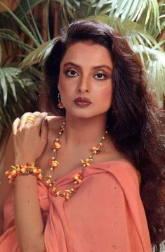 a woman with long brown hair wearing a necklace and dress posing for a photo in front of palm trees