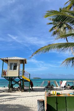 a lifeguard tower on the beach with people sitting and standing around it, next to palm trees